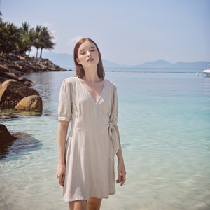 May include: A woman wearing a light beige wrap dress with a V-neckline and short sleeves stands on a beach with the ocean in the background.
