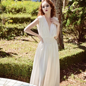 May include: A cream-colored, floor-length dress with a deep V-neck and cap sleeves. The dress features a fitted, crocheted waist and a flowing skirt. The model is standing outdoors in a garden setting, with sunlight filtering through the trees.