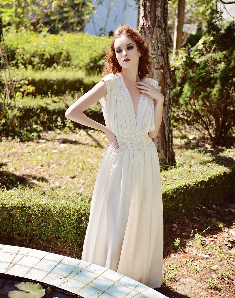 May include: A cream-colored, floor-length dress with a deep V-neck and cap sleeves. The dress features a fitted, crocheted waist and a flowing skirt. The model is standing outdoors in a garden setting, with sunlight filtering through the trees.