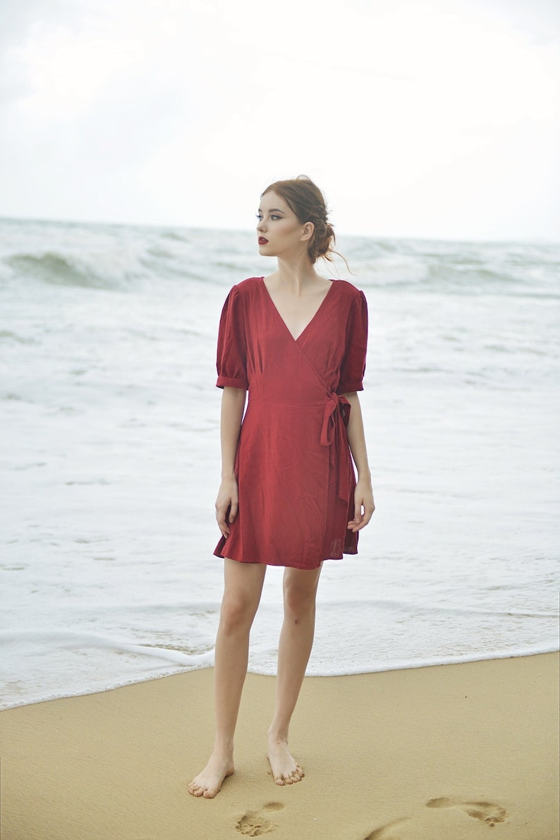 May include: A woman in a red wrap dress stands on a sandy beach with the ocean behind her.