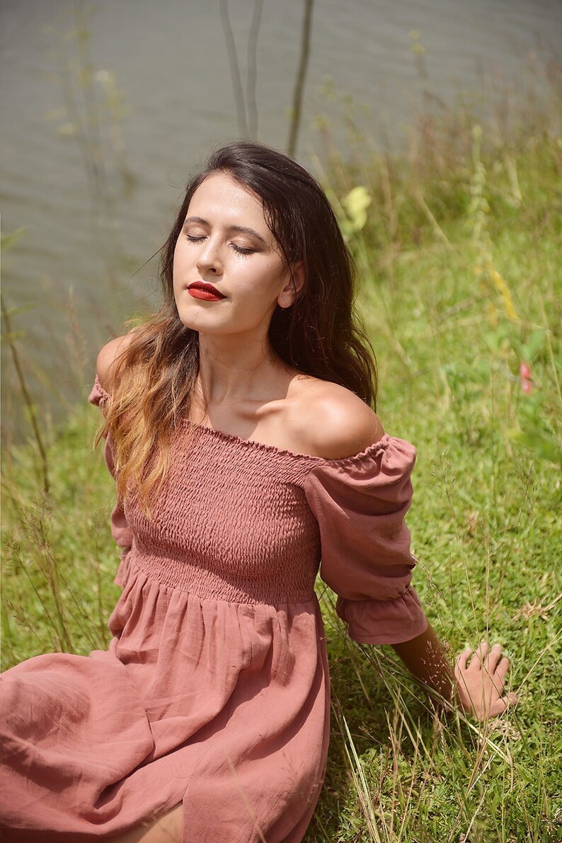 May include: A woman wearing a dusty rose off-the-shoulder dress with puffed sleeves, sitting in a field of green grass.