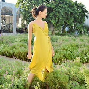 May include: A yellow wrap dress with a V-neck and ruffled trim. The dress is made of a flowing fabric and is worn outdoors in a garden setting. The dress is knee-length and has a tie closure at the waist.