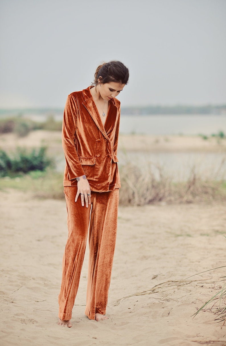 May include: A woman wearing a rust-colored velvet suit with a double-breasted blazer and wide-leg pants. She is standing on a sandy beach with tall grasses in the background.