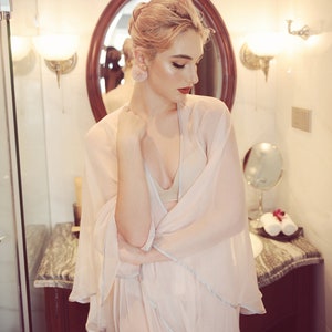 May include: A woman with pink hair is wearing a sheer, light pink robe with a white trim. She is standing in a bathroom with a marble countertop and a mirror behind her.