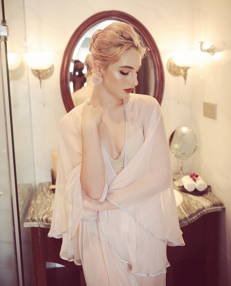 May include: A woman with pink hair is wearing a sheer, light pink robe with a white trim. She is standing in a bathroom with a marble countertop and a mirror behind her.