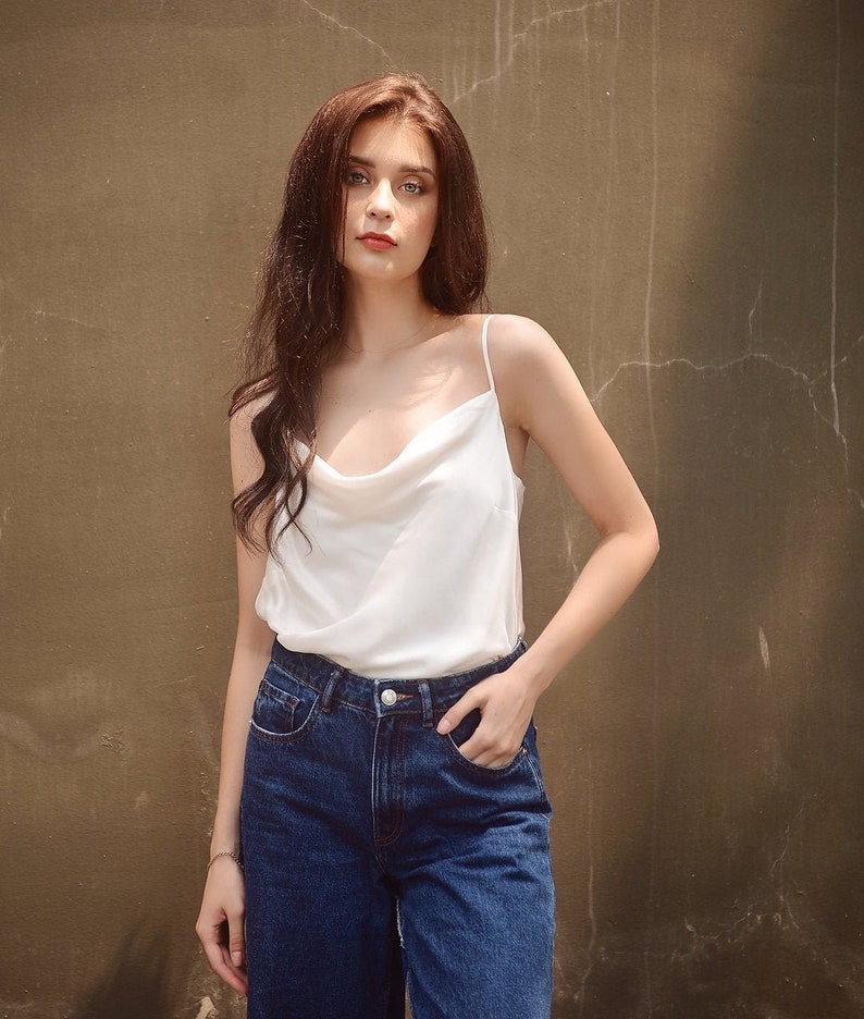 May include: A woman wearing a white cowl neck tank top and blue denim jeans. She is standing in front of a textured brown wall.