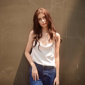 May include: A woman with long brown hair wearing a white tank top and blue jeans stands in front of a brown wall.