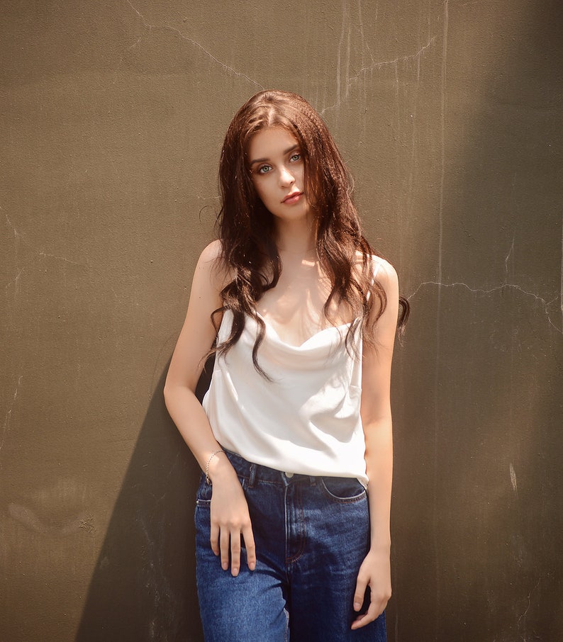 May include: A woman with long brown hair wearing a white tank top and blue jeans stands in front of a brown wall.