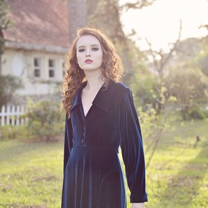 May include: A long, dark blue velvet dress with a collared neckline and long sleeves. The dress has a button-down front and a fitted bodice, with a flowing skirt. The model has long, curly auburn hair.