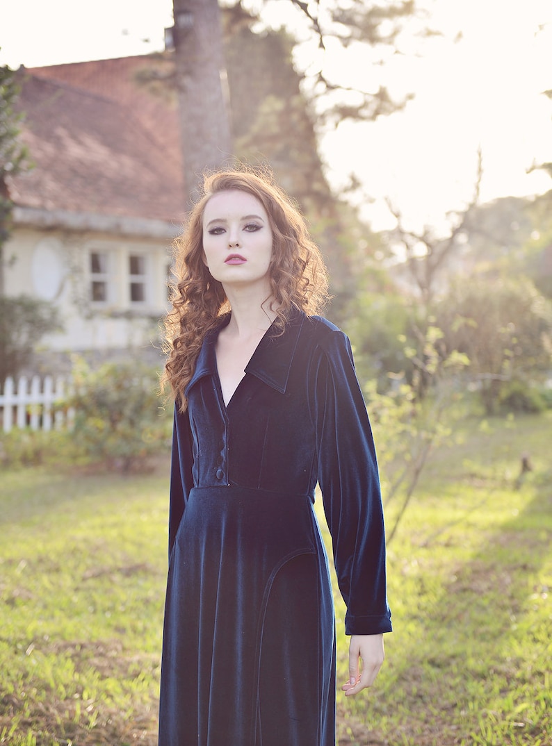 May include: A long, dark blue velvet dress with a collared neckline and long sleeves. The dress has a button-down front and a fitted bodice, with a flowing skirt. The model has long, curly auburn hair.