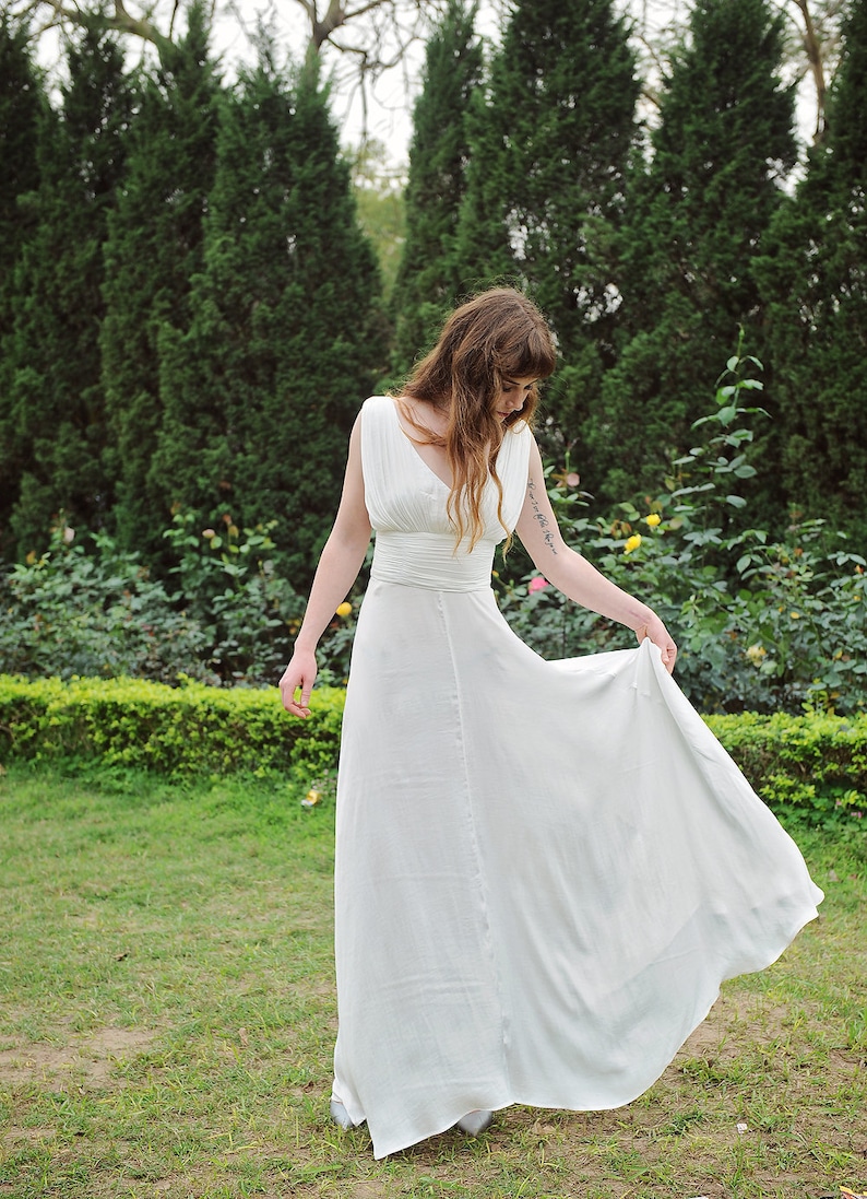 May include: A woman in a white sleeveless maxi dress with a fitted bodice and a flowing skirt. She is standing in a grassy area with a row of evergreen trees in the background.