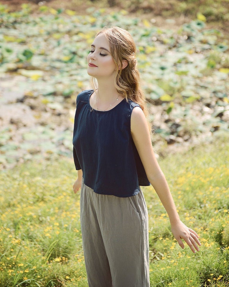 May include: A woman wearing a navy blue sleeveless top and gray pants stands in a field of yellow flowers. She has her eyes closed and is smiling.
