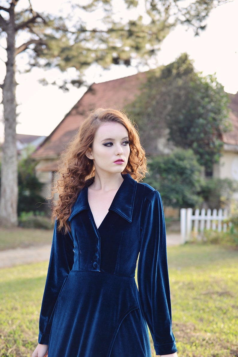 May include: A long-sleeved, dark teal velvet dress with a collared neckline and button details. The dress has a fitted bodice and a flowing skirt. The model is standing outdoors in front of a house and a white picket fence.