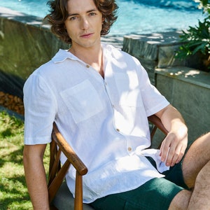 May include: A man wearing a white linen shirt with a chest pocket and green linen shorts sits on a wooden chair in a grassy area.