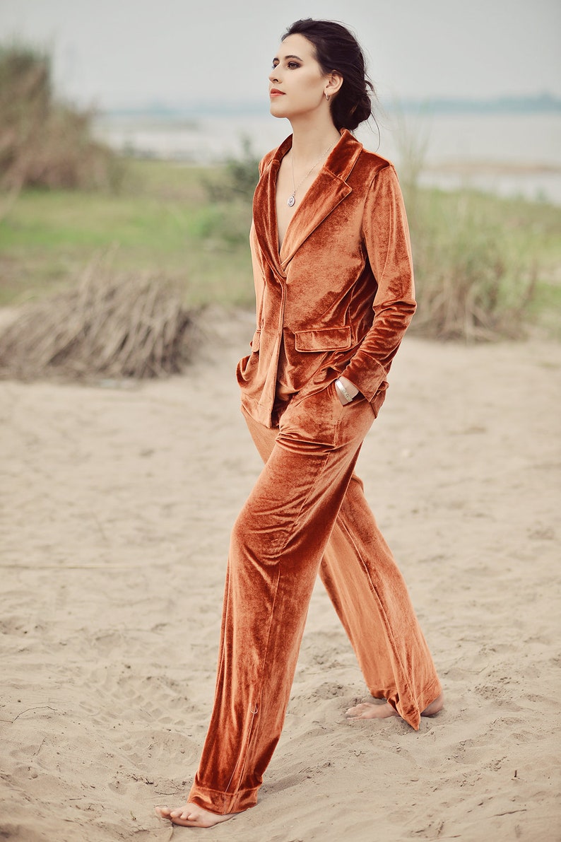 May include: A woman wearing a rust-colored velvet suit with a long-sleeved blazer and wide-leg pants. She is standing on a sandy beach with her hands in her pockets.