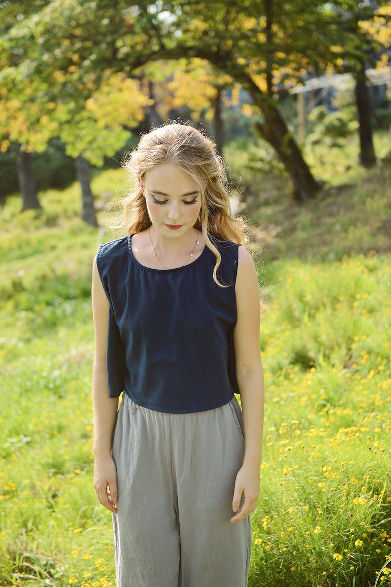 May include: A woman wearing a dark blue sleeveless top and light gray pants stands in a field of yellow wildflowers. She has long blonde hair and is looking down.