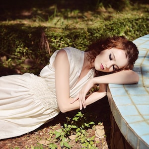 May include: A woman wearing a cream-colored dress lies near a light blue tiled structure. Her red hair is styled in loose curls. The dress has a V-neck and a gathered waist. The scene is set outdoors with greenery in the background.