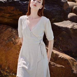 May include: A woman with red hair wearing a light beige wrap dress with short sleeves and a tie at the waist. She is standing in front of large rocks.