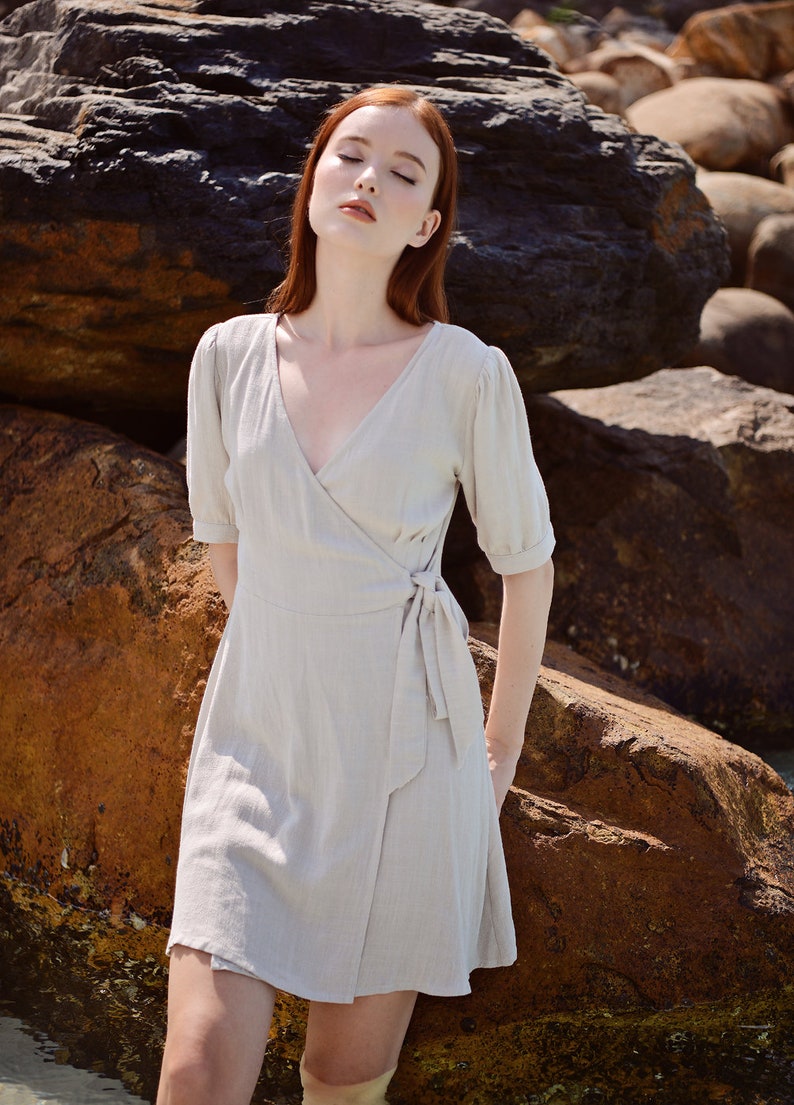 May include: A woman with red hair wearing a light beige wrap dress with short sleeves and a tie at the waist. She is standing in front of large rocks.