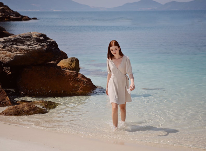 May include: A woman in a light beige linen dress walks along a sandy beach with clear blue water. She is walking towards the camera with her head down.