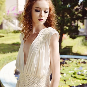 May include: Ivory-colored sleeveless dress with a deep V-neckline and a fitted waist. The dress features delicate smocking details on the bodice and waist. The fabric appears lightweight and flowing, suitable for warm weather. The dress is a classic, elegant design.
