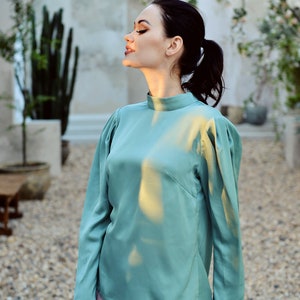 May include: A woman wearing a teal long-sleeved blouse with a high neckline and a pink skirt.