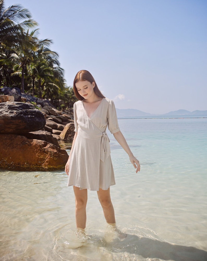 May include: A woman wearing a light beige wrap dress stands in shallow water on a beach. The water is clear and the sand is white. The woman is looking down at the water.
