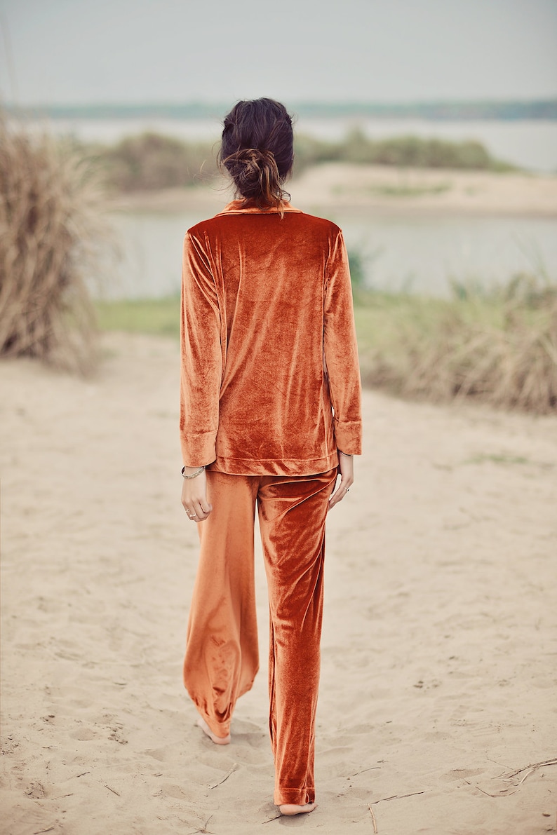 May include: A person wearing a rust-colored velvet pajama set with long sleeves and wide-leg pants. The person is walking on a sandy beach.