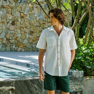 May include: A white linen shirt with a button-down collar and short sleeves. The shirt has a chest pocket and is worn with dark green shorts.