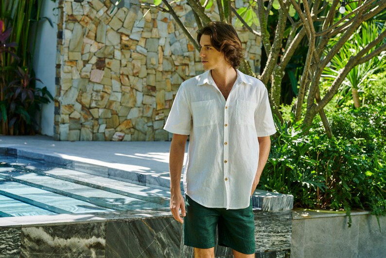 May include: A white linen shirt with a button-down collar and short sleeves. The shirt has a chest pocket and is worn with dark green shorts.