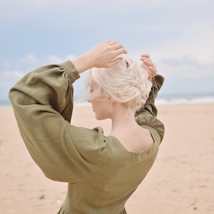 May include: A woman with blonde hair wearing a long-sleeved, olive green dress with puffed sleeves. She is standing on a sandy beach with the ocean in the background.