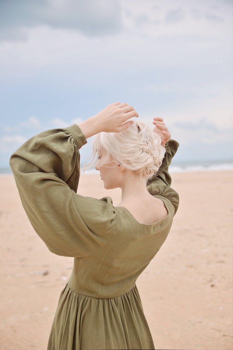May include: A woman with blonde hair wearing a long-sleeved, olive green dress with puffed sleeves. She is standing on a sandy beach with the ocean in the background.