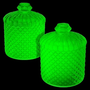 May include: Two bright green glass jars with lids. The jars have a diamond pattern and a ribbed design on the lids. The jars are glowing against a black background. The jars are likely vintage or antique.