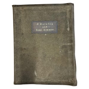 Puede incluir: Un libro rectangular y desgastado con una cubierta verde oscuro. Una etiqueta rectangular en la cubierta dice "a Catalog and Some Comment". La cubierta del libro muestra signos de desgaste y envejecimiento.