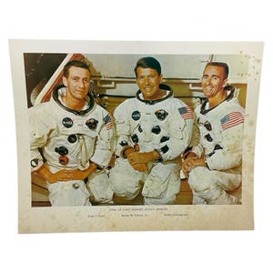 Puede incluir: Fotografía vintage con tres astronautas con trajes espaciales blancos, cada uno con un parche de la NASA y una bandera estadounidense. La imagen está etiquetada como "CREW OF FIRST MANNED APOLLO MISSION" con los nombres Donn F. Eisele, Walter M. Schirra, Jr. y Walter Cunningham.