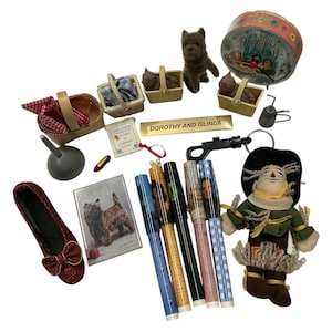 May include: A collection of Wizard of Oz themed items, including a red glitter shoe, a dog figurine, baskets, pens, a tin, and a scarecrow doll. The items are arranged on a white surface.