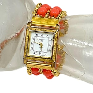 Puede incluir: Reloj cuadrado dorado con una esfera blanca y marcadores de hora en números romanos. El reloj tiene una pulsera de eslabones con cuentas de color coral.