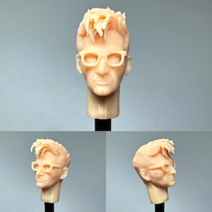 Könnte beinhalten: Detaillierte Skulptur eines menschlichen Kopfes mit kurzen, stacheligen Haaren und Brille. Der Kopf ist hellbeige und auf einem schwarzen Ständer montiert. Die Brille hat einen rechteckigen Rahmen. Die Skulptur ist realistisch.