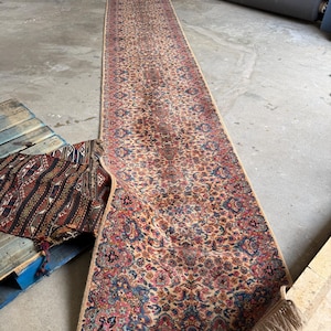Pode incluir: Um tapete longo e retangular com um padrão floral detalhado em tons de bege, vermelho, azul e rosa. O tapete tem uma borda bege e franjas nas extremidades. O tapete está parcialmente enrolado, revelando seu comprimento e design intrincado.