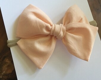 May include: A peach-colored fabric bow on a light green headband, resting on a white card. The card reads "BOWS BY MADILYN NICOLE" at the top and "HANDMADE FOR YOU" at the bottom. The bow is centered and the background is a wooden surface.