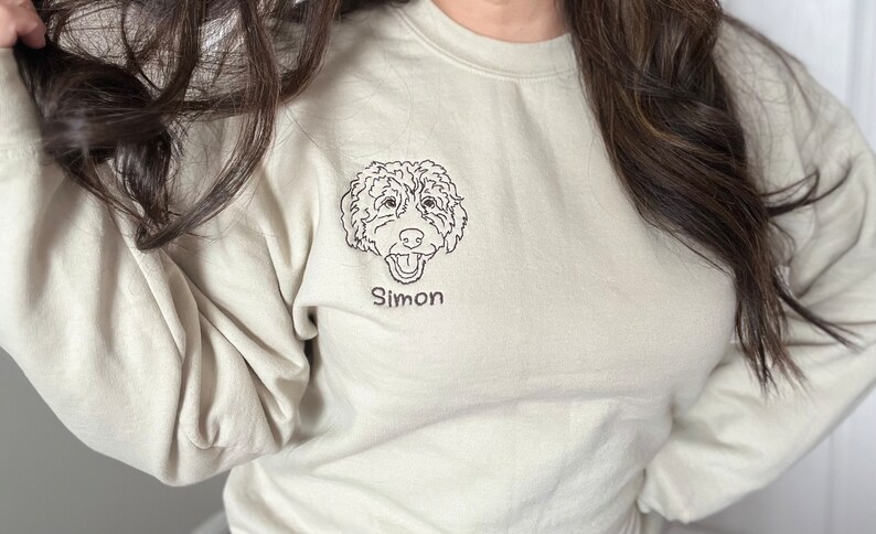 May include: A beige sweatshirt with a black line drawing of a dog's head and the name "Simon" embroidered on the front.
