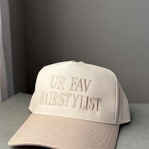 May include: A beige baseball cap with a curved peak. The cap has the embroidered text "UR FAV HAIRSTYLIST" in a matching beige thread. The hat is made of a sturdy fabric and has a classic six-panel design.