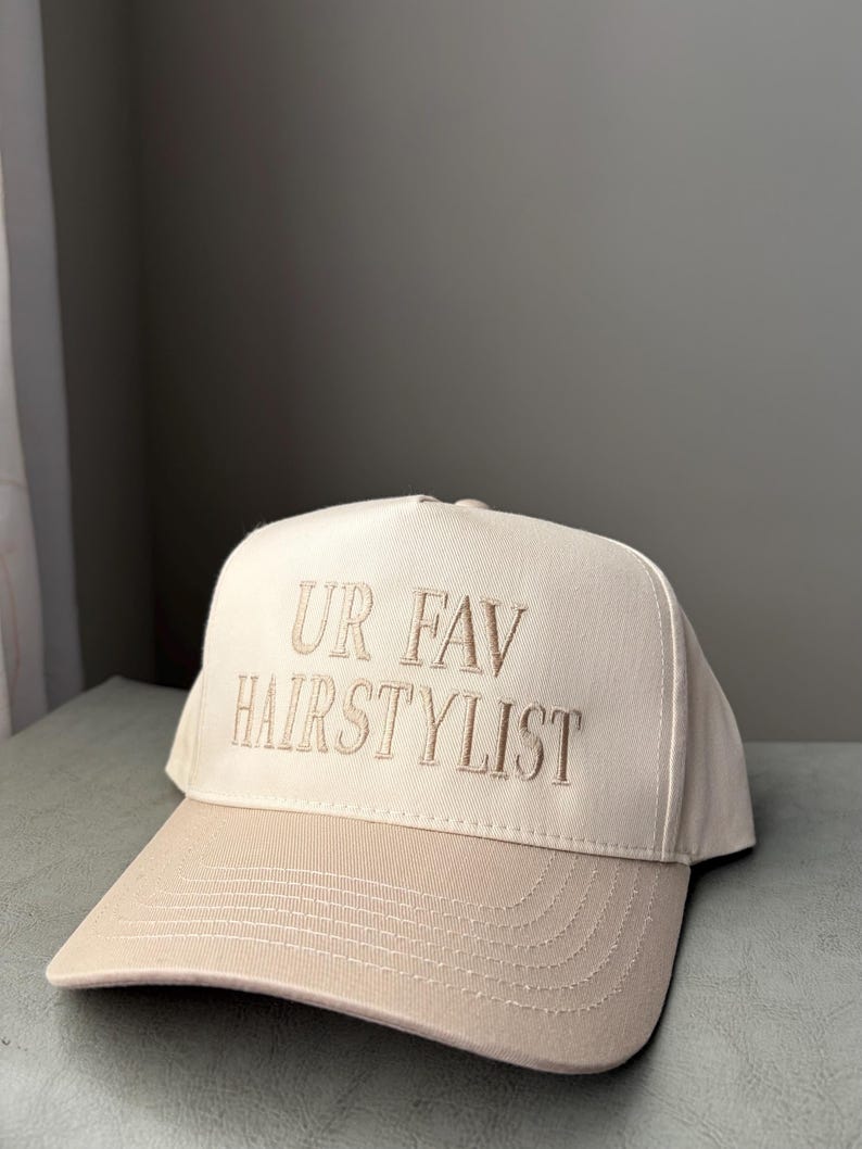 May include: A beige baseball cap with a curved peak. The cap has the embroidered text "UR FAV HAIRSTYLIST" in a matching beige thread. The hat is made of a sturdy fabric and has a classic six-panel design.