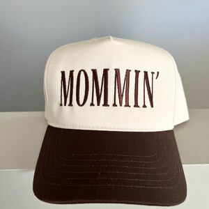 Puede incluir: Gorra de b&eacute;isbol blanca con visera marr&oacute;n. La palabra "MOMMIN'" est&aacute; bordada en marr&oacute;n en la parte delantera. La gorra est&aacute; hecha de un material resistente y tiene un estilo cl&aacute;sico e informal.