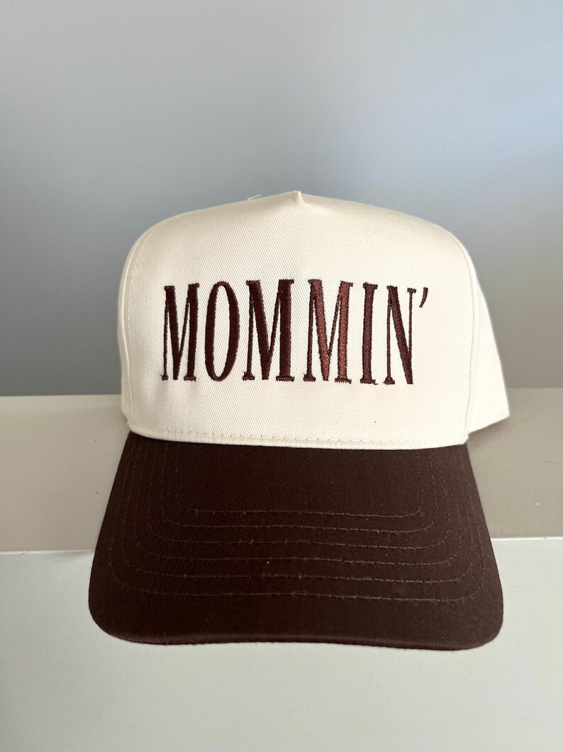 Puede incluir: Gorra de b&eacute;isbol blanca con visera marr&oacute;n. La palabra "MOMMIN'" est&aacute; bordada en marr&oacute;n en la parte delantera. La gorra est&aacute; hecha de un material resistente y tiene un estilo cl&aacute;sico e informal.
