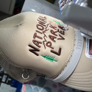 May include: A beige trucker hat with brown embroidery that reads "National Park Lover" with a green pine tree and mountain graphic.