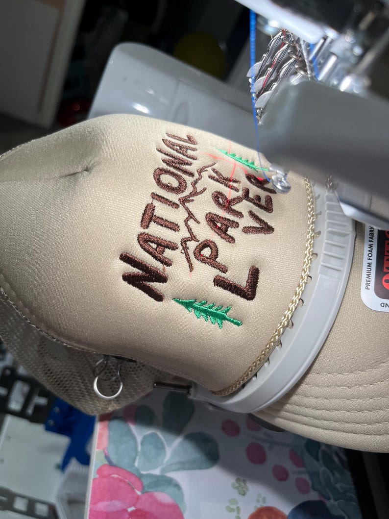 May include: A beige trucker hat with brown embroidery that reads "National Park Lover" with a green pine tree and mountain graphic.
