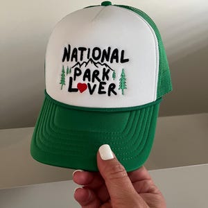 May include: White and green trucker hat with the text "NATIONAL PARK LOVER" embroidered in black with a red heart. The hat has a green mesh back.