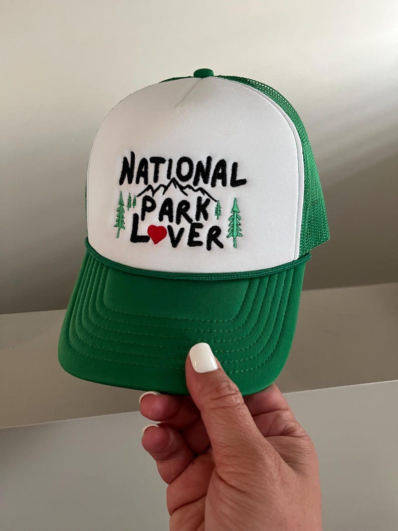 May include: White and green trucker hat with the text "NATIONAL PARK LOVER" embroidered in black with a red heart. The hat has a green mesh back.
