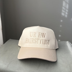 May include: A cream-coloured baseball cap with a tan brim. The phrase "UR FAV HAIRSTYLIST" is embroidered in a matching tan thread across the front of the cap. The cap is sitting on a light blue surface.